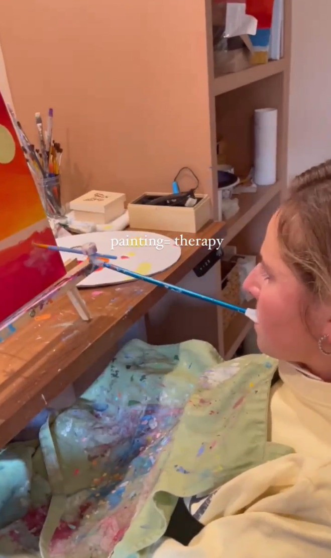 A screenshot of a vertical video of Rebecca mouth-painting a vivid sunrise or sunset scene in orange, red, and yellow. She wears a canvas piece to prevent splatters that has lots of colorful paint on it, and she has light skin and blonde-brown hair pulled back.