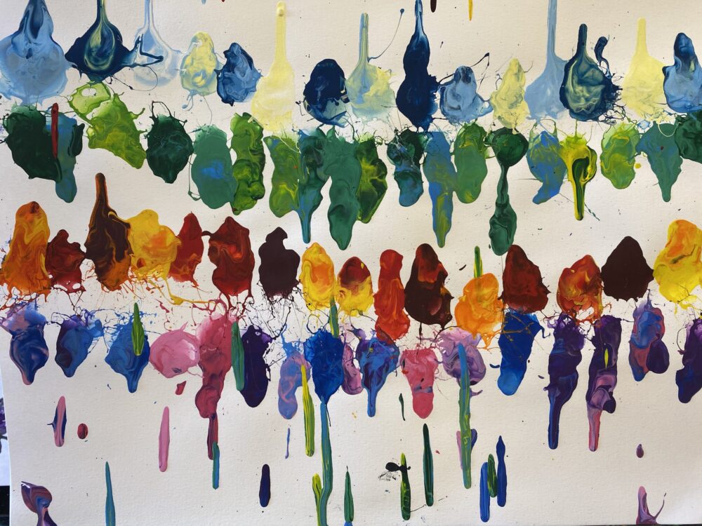 A piece of art made by Rebecca Koltun with a straw blowing technique features artistically arranged paint blobs in multiple eye-catching shades of blue-green, yellow-green, dark red, orange, purple, and blue in four lines with a drip at the top and bottom.