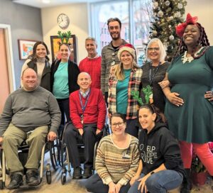 Members of the Help Hope Live staff and board with staff at Magee Rehabilitation in the Magee Rehabilitation facility with windows showing a city street and red brick buildings and a decorated holiday tree with silver ornaments. The individuals pictured are a diverse mix of ages and genders with most standing and two individuals seated in their wheelchairs. Many wear red, green, or other festive holiday touches.