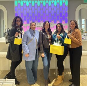 At a Kendra Scott store, five Help Hope Live staff and board members hold up their bright yellow Kendra Scott shopping bags. They are all women of various ages and ethnicities in business-casual winter wear.