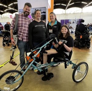 Kelly L Green is with 15-year-old Hallie and her parents at Abilities Expo Dallas. Hallie is sitting on her new teal Help Hope Live-themed handcycle bike. She has light skin, shoulder-length brown hair, a black t-shirt, and one leg. Her dad has light skin, thick gray hair, and a gray short beard. He wears jeans and a button-up shirt and carries Hallie’s prosthetic leg, which ends in a hiking boot. Her mom has light skin, brown hair pulled back, and a black athletic long-sleeved shirt. Kelly has light skin and dark hair in a ponytail with a black Help Hope Live jacket and teal polo collar.