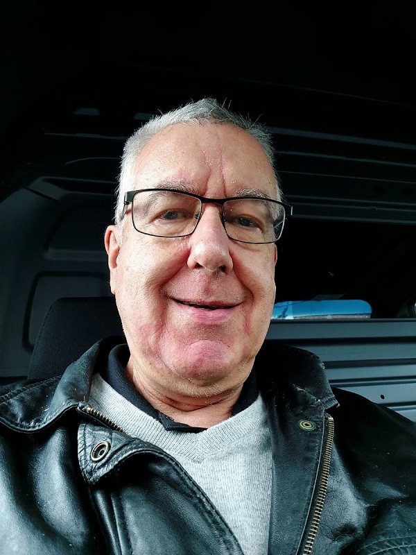 In a car selfie, heart transplant recipient Greg Wright has light skin, short gray hair, light eyes, and black-rimmed glasses with a worn black leather jacket over a gray sweater.