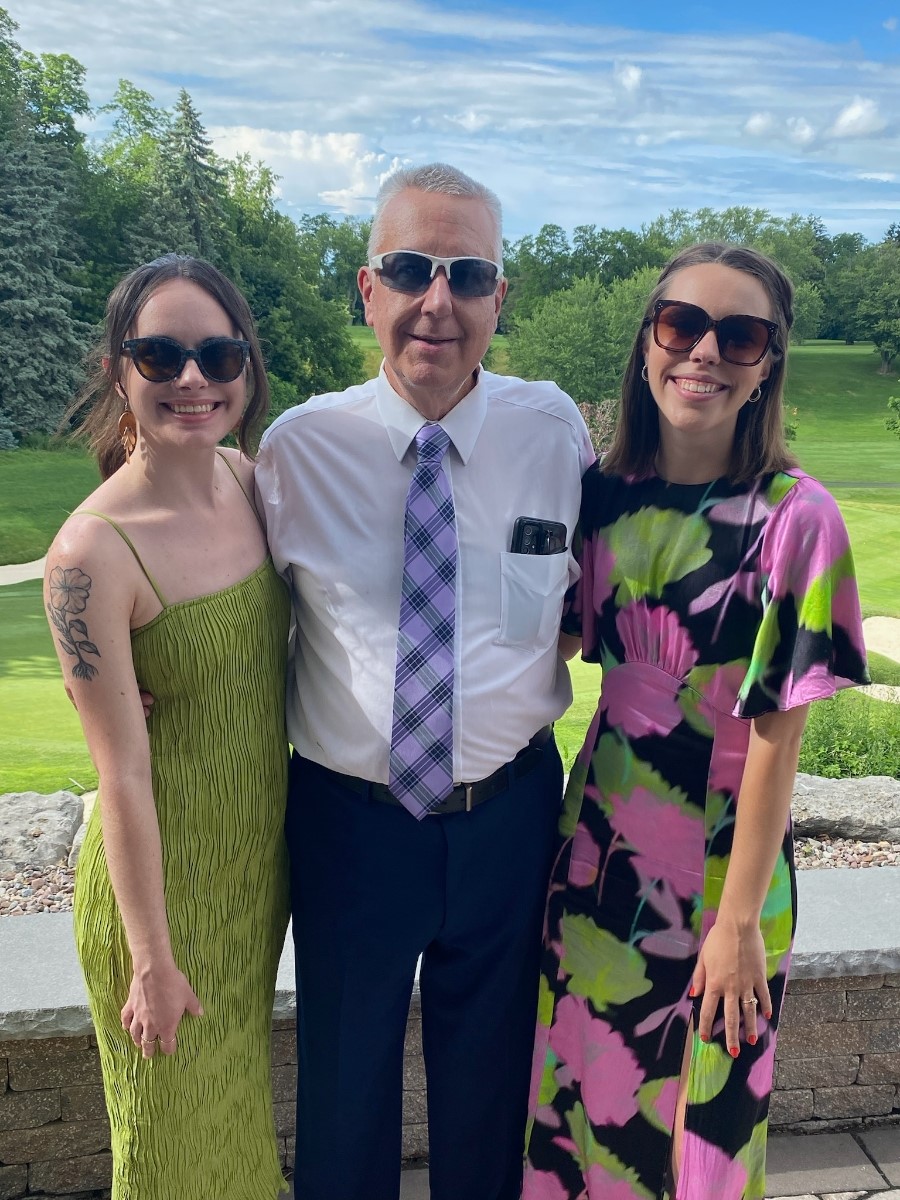 Heart transplant recipient Greg Wright with his daughters Jessica and Megan. Greg has light skin, short gray hair, sunglasses, and formal attire. Jessica has light skin, brown hair pulled back, sunglasses, and a spring green sundress. Megan has light skin, shoulder-length straight brown hair, sunglasses, and a graphic flower print dress with vivid green and pink designs on black. They are outdoors near a golf course setting.