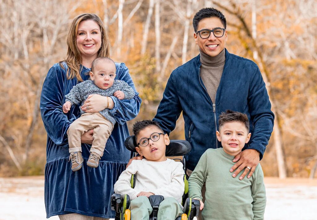 Oliver Garcia with his family in a portrait taken outdoors with fall trees visible in the background. Oliver is a boy with light brown skin, glasses, and short dark hair seated in his wheelchair. His brother beside him also has short dark hair and light brown skin and is standing. Oliver's mother is holding a baby and has light skin, shoulder-length blonde hair, and a blue velvet dress. His dad has light brown skin, glasses, and short dark hair and wears a turtleneck and blue velvet jacket.