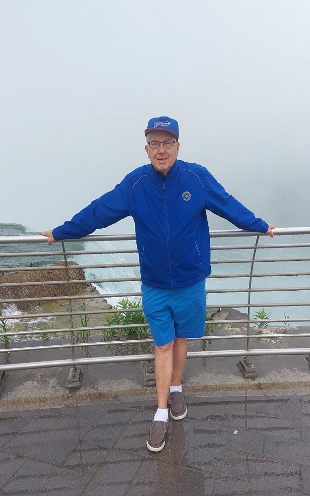 Pictured in front of a railing atop Niagara Falls, heart transplant recipient Greg Wright has light skin, short gray hair, glasses, a deep blue windbreaker, and blue shorts with a blue baseball cap.