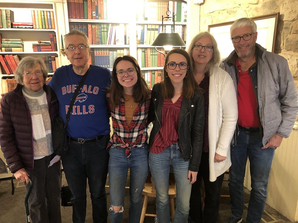 Heart transplant recipient Greg Wright with his two daughters and three other members of his family in a home library setting. Greg has light skin, short gray hair, a navy Buffalo Bills long-sleeved shirt, and black jeans. His daughters both have light skin, dark eyes, straight dark hair, and glasses.