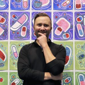 Transplant recipient and artist Dylan Mortimer has light skin, short brown hair, and a short brown beard. He is raising his left hand thoughtfully to his chin. Behind him is tiled artwork by him depicting pills and abstract antibody-like shapes in pink, white, red, blue, and green with glitter.