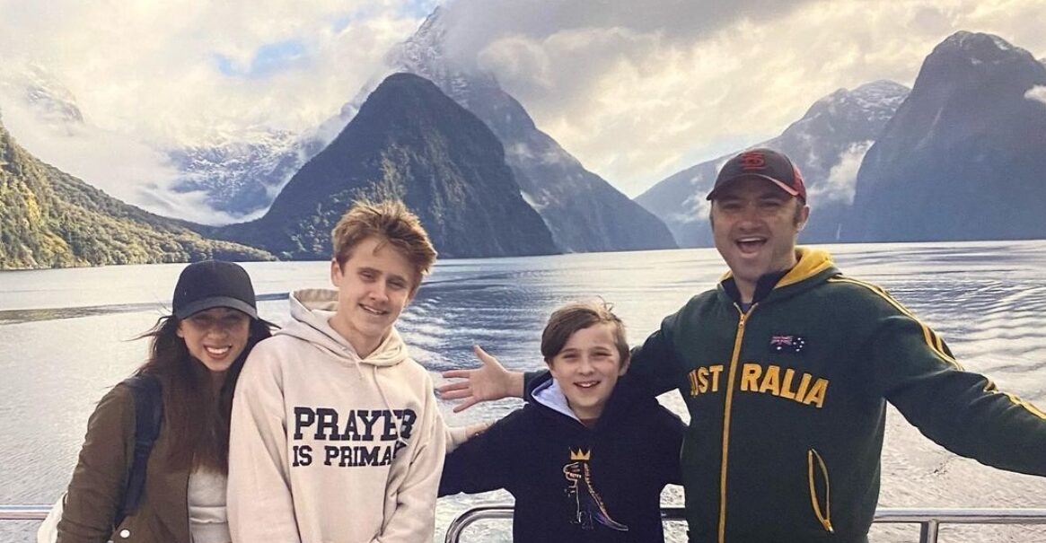 Transplant recipient and artist Dylan Mortimer has light skin, short brown hair, and a short brown beard as he stands in front of a gorgeous New Zealand lake and mountain landscape with three members of his family. From left, there is a young woman with light skin, brown hair past her shoulders, and a navy baseball cap, then a teen with light skin, short curly brown hair, and a cream sweatshirt with the word PRAYER visible in navy print, then a younger boy with short straight brown hair and light skin, then Dylan wearing an AUSTRALIA printed hoodie.