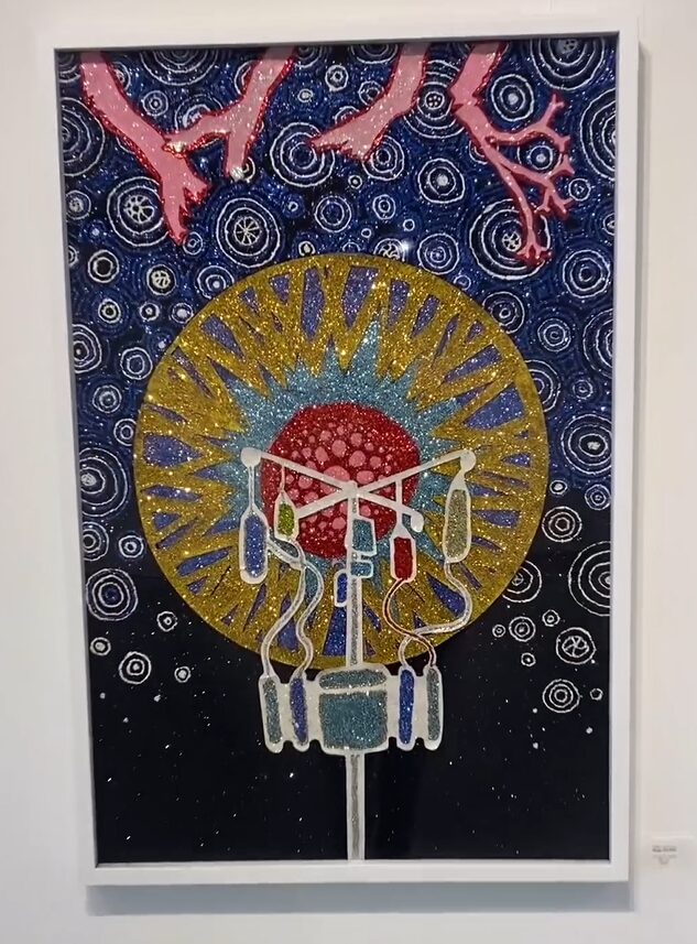 An artwork by Dylan Mortimer hanging on a wall in a white frame with a gallery or museum placard beside it, not legible. The piece of art features an IV cart in front of a blooming, mandala-like display reminiscent of both anatomy and nature. There is a red globule in the center surrounded by a turquoise and glittering gold circular halo. Behind that, a deep blue fading to black near the bottom of the canvas offsets silvery glitter swirls and pink protuberances that look like arteries or ventricles.