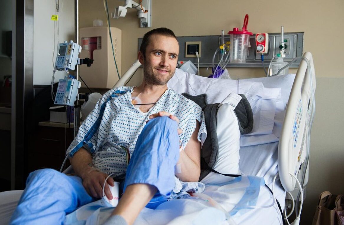 Seated in a hospital bed wearing a white and blue hospital gown, transplant recipient and artist Dylan Mortimer has light skin, short brown hair, and a short brown beard.