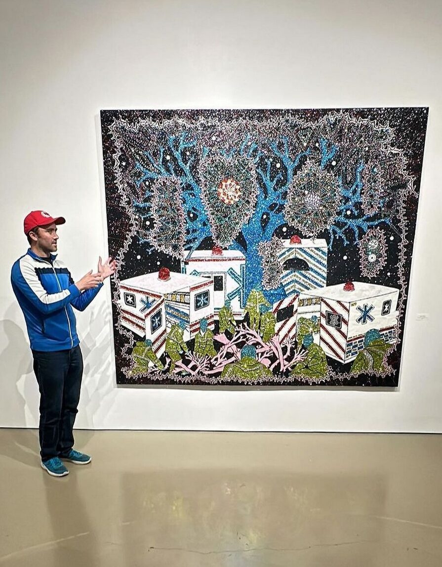 Transplant recipient and artist Dylan Mortimer has light skin, short brown hair, and a short brown beard and he wears a red cap and sneakers as he addresses a non-visible crowd in front of one of his pieces of art hanging in a gallery or museum setting. The work of art is taller and longer than Dylan's height and features a scene of ambulances and snaking, flora-like ventricle and artery shapes that bring a set of lungs to mind. His works often feature glitter.