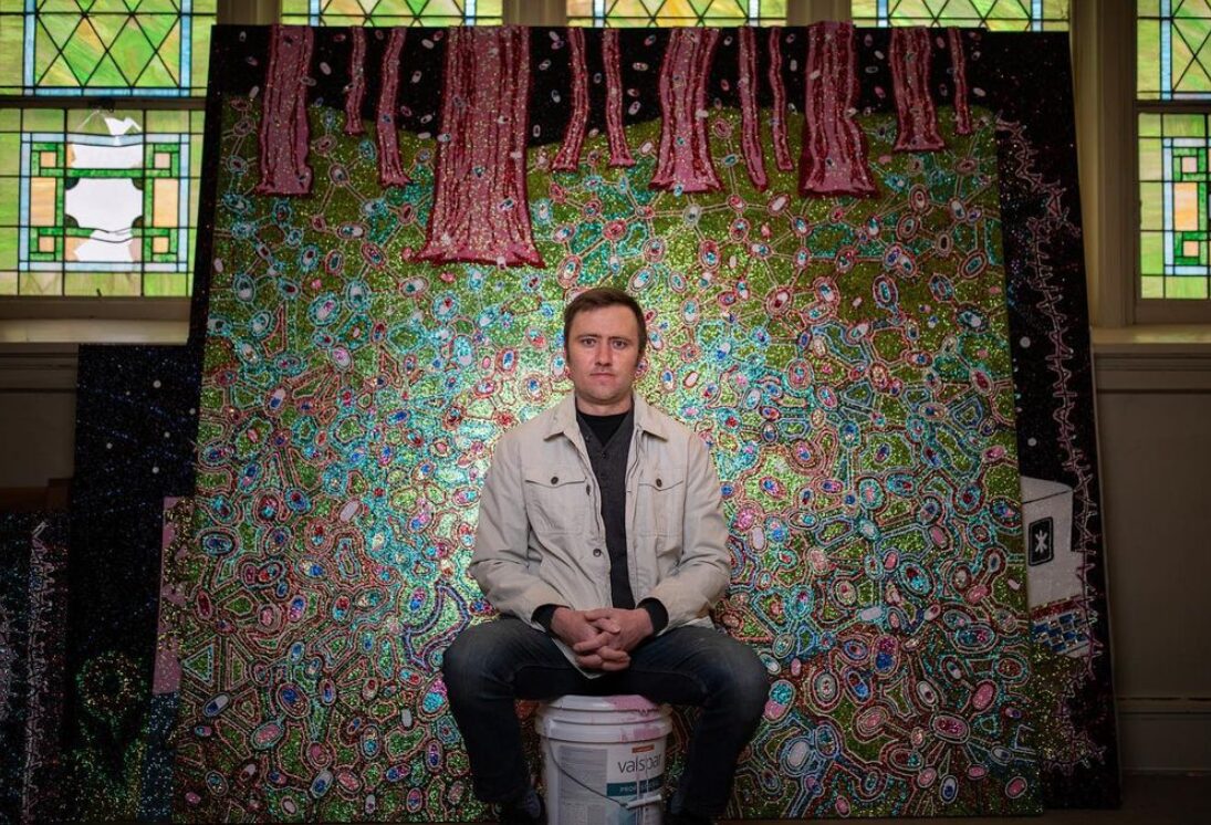 Transplant recipient and artist Dylan Mortimer has light skin and short brown hair and wears jeans and a white pocketed jacket as he sits on a paint can with a spotlight on him in front of one of his artworks. The work appears to be about as tall and wide as Dylan is tall. It features red and pink tree-like but anatomical structures blooming from a 'forest floor' of jewel-toned and glitter-accented amoeba-like shapes.