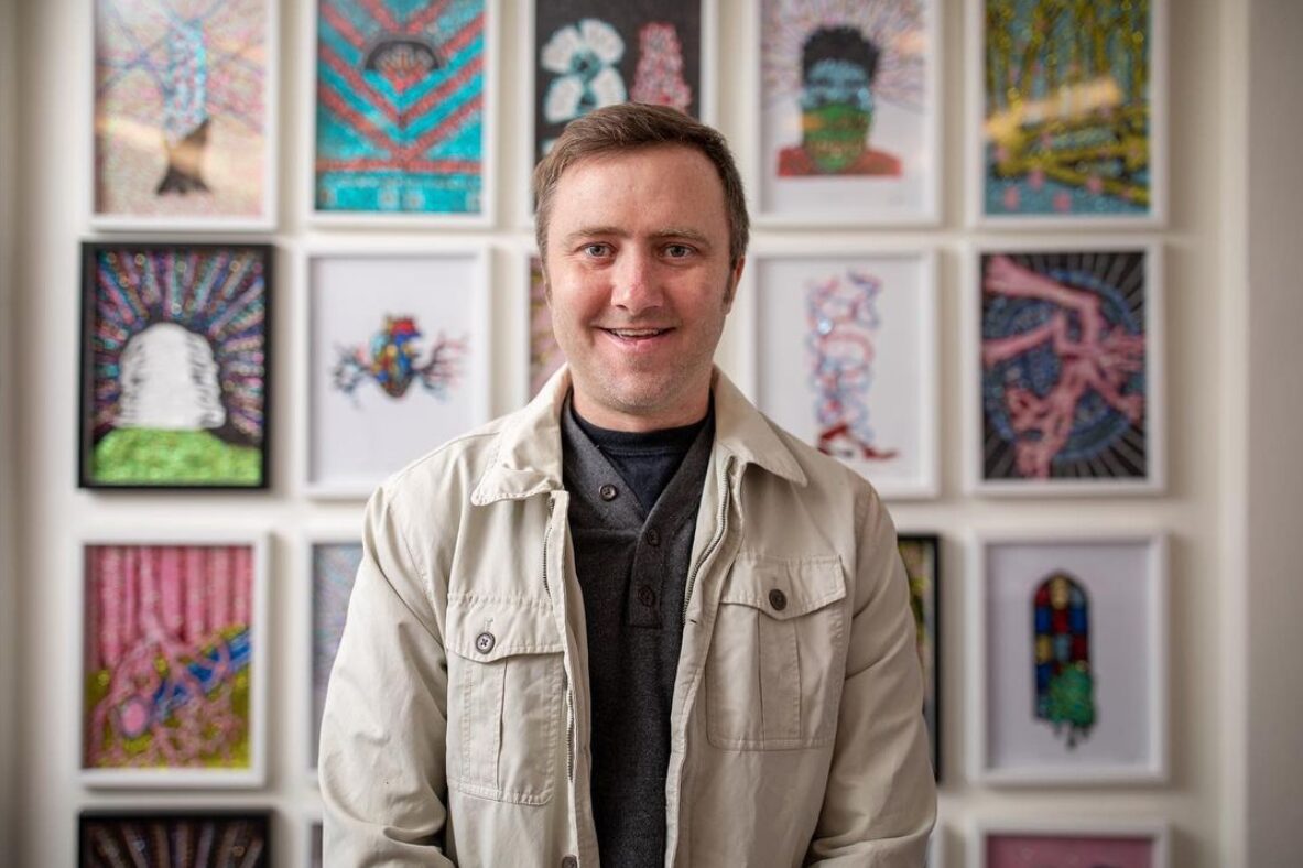 Transplant recipient and artist Dylan Mortimer has light skin and short brown hair and wears a navy shirt and a cream cargo jacket as he stands in front of an assortment of small framed artworks by him, featuring jewel toned colors and glitter.