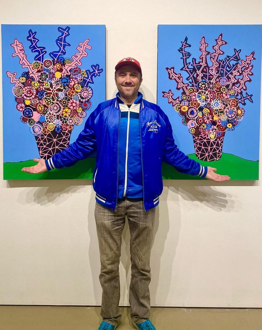 Transplant recipient and artist Dylan Mortimer has light skin, short brown hair, and a short brown beard and he wears a red baseball cap and a vivid deep cerulean jacket as he stands in a gallery or museum setting in front of two of his artworks. The artworks are about three-quarters of Dylan's height in size, and they feature blooming bouquets of surprisingly anatomical designs like neural pathways in vivid jewel tones of pink, red, orange, yellow, and blue.