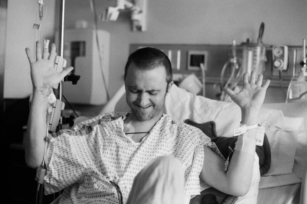 In a powerful image in black and white, transplant recipient and artist Dylan Mortimer has light skin, short shaved brown hair, and a short brown beard and he raises both his hands in a worship-like gesture as he sits in a hospital bed wearing a hospital gown. His chin is lowered with his eyes closed and his eyebrows wrinkled with concentration or emotion.