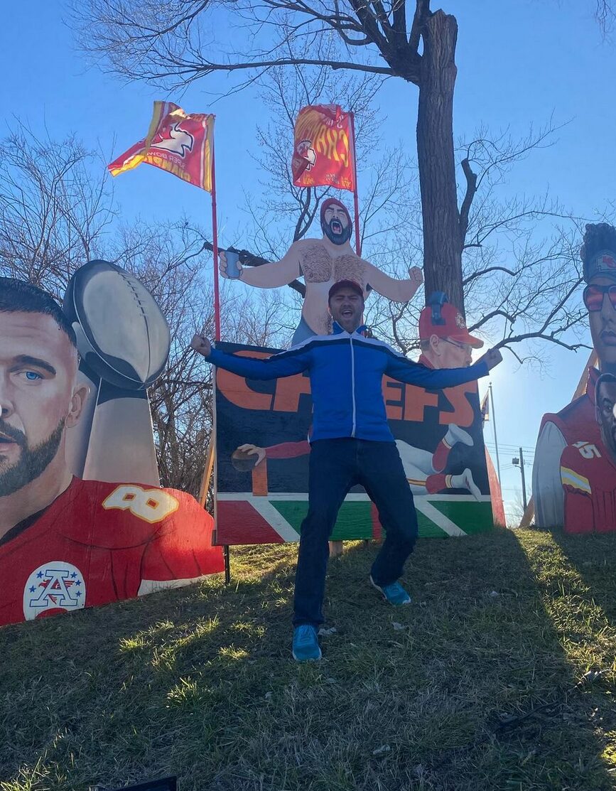 Transplant recipient and artist Dylan Mortimer has light skin, short brown hair, and a short brown beard and wears a red cap and a bright vivid cerulean jacket and sneakers. He is on a lawn outdoors replicating a fan-made artwork behind him of NFL player Jason Kelce shirtless and supporting his brother's Kansas City Chiefs team during their 2024 Super Bowl bid. There is additional large artwork of Travis Kelce and Patrick Mahomes, Chiefs NFL players, in their signature red and white jerseys.