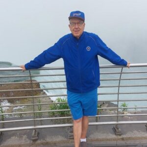 Pictured in front of a railing atop Niagara Falls, heart transplant recipient Greg Wright has light skin, short gray hair, glasses, a deep blue windbreaker, and blue shorts with a blue baseball cap.