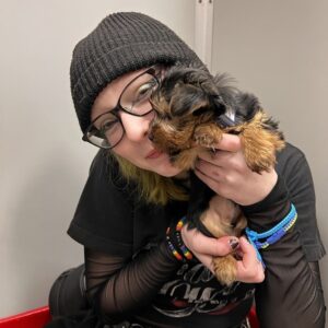 Teenager Aislyn Moss holds a small terrier up to her cheek and smiles. She has light skin, black-framed glasses, gold eyeliner, a gray beanie, a black t-shirt with full-length mesh sleeves, and some rainbow beaded bracelets.