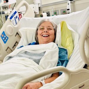 Transplant recipient Valerie is beaming as she lies in a hospital bed wearing a blue and green print hospital gown. She has light skin, clear-rimmed glasses, and straight gray hair.