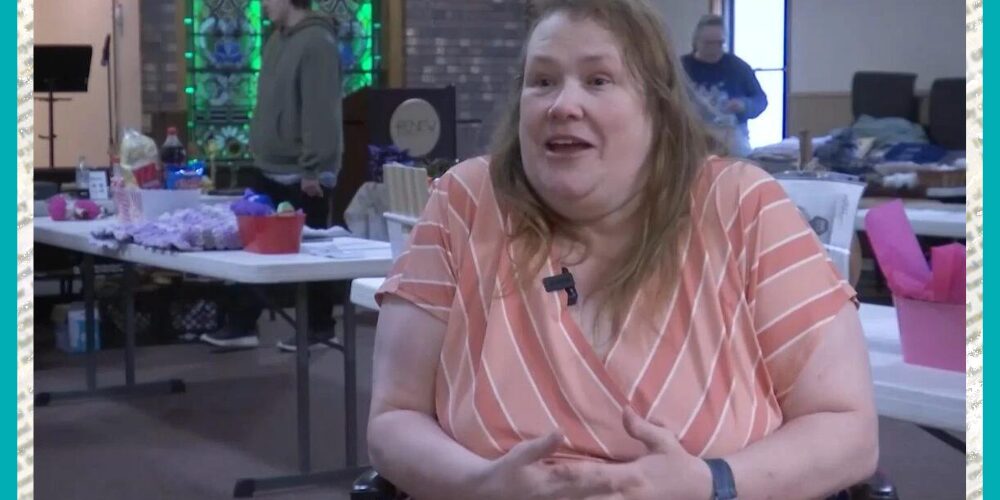 Speaking excitedly to a news team in a news broadcast screenshot, client Lisa Marie Lee is seated in her wheelchair with a raffle or auction fundraiser scene visible behind her. She has light skin, brown hair past her shoulders, dark eyes, and a salmon pink striped dress with short sleeves and a V-neck.