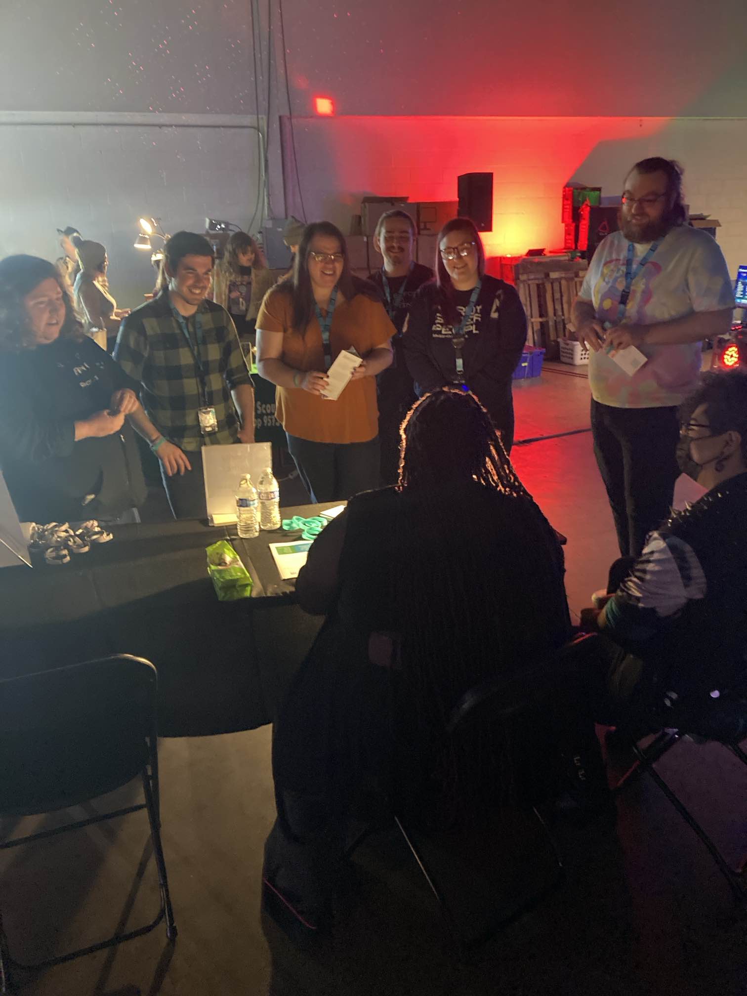 In a photo taken from behind charity rep Vanessa's head as she is seated at a table, six excited gamers grin as they learn about Help Hope Live at the charity table of RGB LAN 6. They are various ages and genders. Vanessa's long dreadlocks are visible and her partner's brown skin and short curly black hair and black face mask.