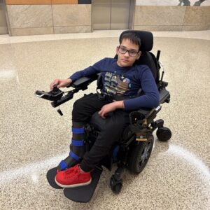 12-year-old Milen Kichukov is pictured seated in his black power chair. He has light skin, short dark hair, glasses, a navy long-sleeved shirt, and bright red sneakers with a leg brace or boot on his right side.