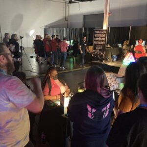 Vanessa Nichols, a Help Hope Live team member, is seated at a Help Hope Live table at the RGB LAN 6 gaming event surrounded by gamers. She has brown skin, red dyed dreadlocks, and a color-blocked pink turtleneck dress in light and dark pink.