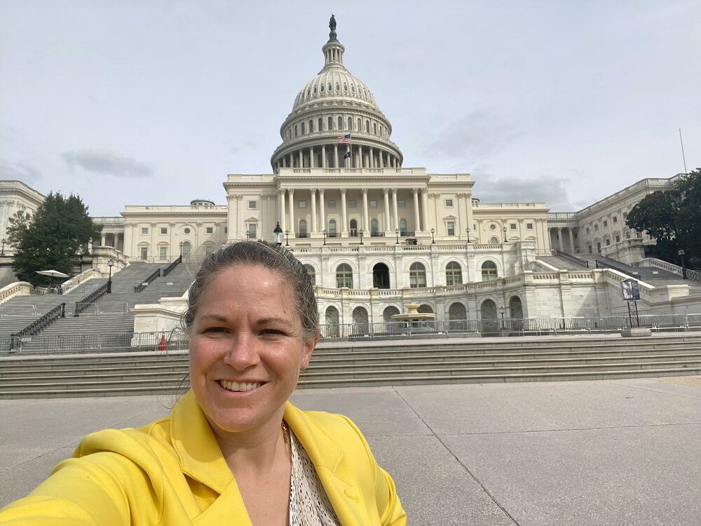 Sonny takes a selfie in front of the capital building in Washington DC. Sonny has light skin, pulled back hair, and a bright lemon yellow blazer.