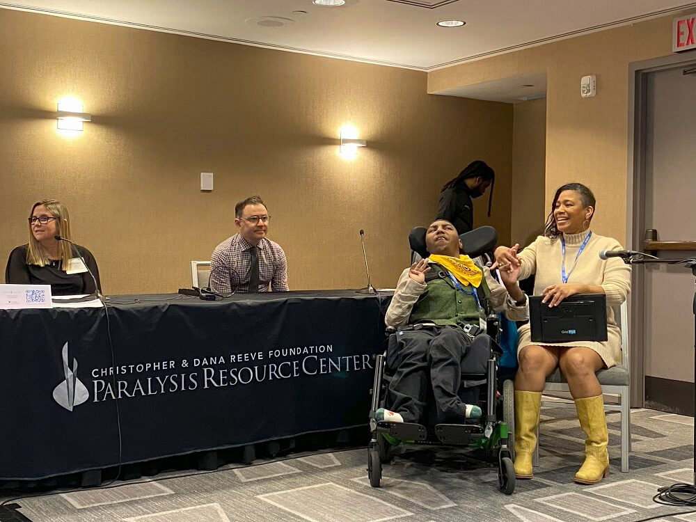 Client Noah Williams and his mom are presenting at Reeve Summit 2024. Noah has brown skin and a bright yellow bandana over a forest green vest. He is seated in a black wheelchair gazing upward. His feet, in socks, point left and right. Mom Naomi has brown skin, a cream colored turtleneck dress, yellow cowboy boots, and a speech communication device on her lap.