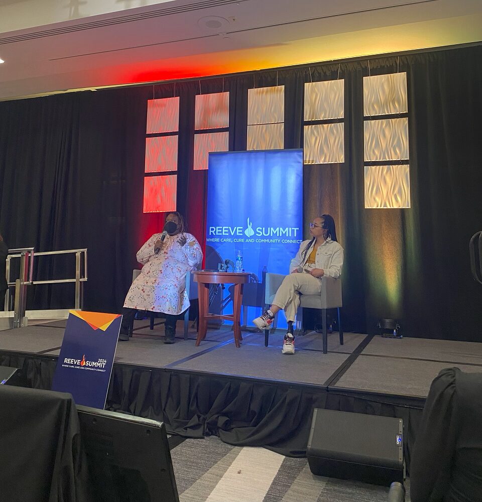 On the presenter stage at Reeve Summit 2024 are two women of color. The first is Imani Barbarin aka Crutches and Spice, who has brown skin, a black face mask, and a floral-printed dress. The second has brown skin, long black braids, a khaki colored jacket, and a prosthetic right leg.