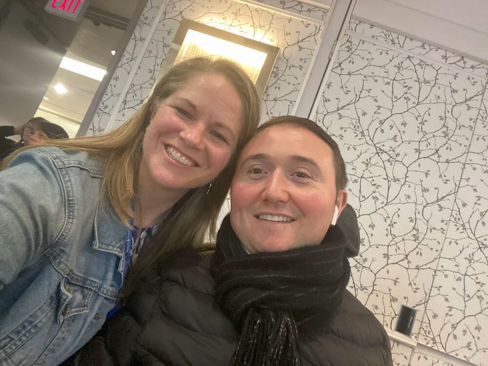 Sonny with Help Hope Live client Ambassador Josh Basile at the Reeve Summit 2024. Sonny has light skin, straight sandy hair past her shoulders, and a denim jacket. Josh has light skin, short brown hair, rosy cheeks, brown eyes, and a black scarf and puffer jacket.