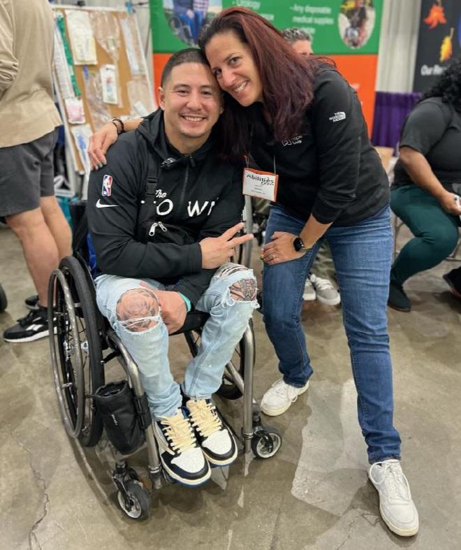 Kelly L Green meets Arthur Renowitzky, founder of Life Goes On Foundation, at Abilities Expo LA 2024. Kelly has light skin, straight red-brown hair, a black Help Hope Live North Face polo, and jeans. Arthur is seated in his black and silver wheelchair with light skin, short black hair, a hoodie, and ripped jeans revealing tattoos on his knees.