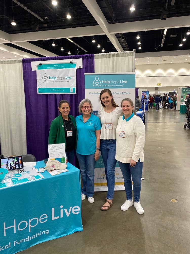 Three Help Hope Live staff members with a client standing at the Help Hope Live branded booth of Abilities Expo 2024. From left to right, Kelly Green has light skin, dark eyes, and an emerald green blazer. Client Ambassador Rachelle Ledbetter has light skin, glasses, shoulder-length gray hair, and a teal Help Hope Live polo. A head taller than the other three women, Karli has light skin, straight brown hair, and a white top. Sonny has light skin and a cream sweater over a teal polo.