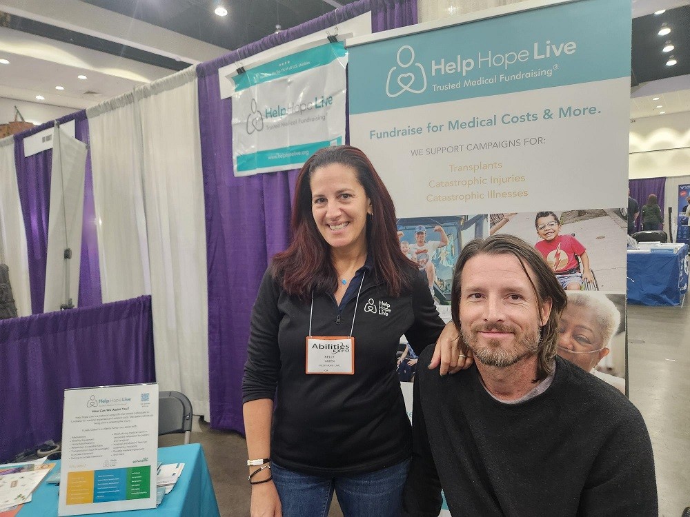 Kelly L Green with client Arthur Martinot in front of the Help Hope Live booth with tagline Trusted Medical Fundraising at Abilities Expo LA 2024. Kelly has light skin, dark eyes, straight red and brown hair, a black Help Hope Live polo sweater, and jeans. Arthur has light skin, shoulder-length brown hair, brown eyes, and a brown and gray short beard.