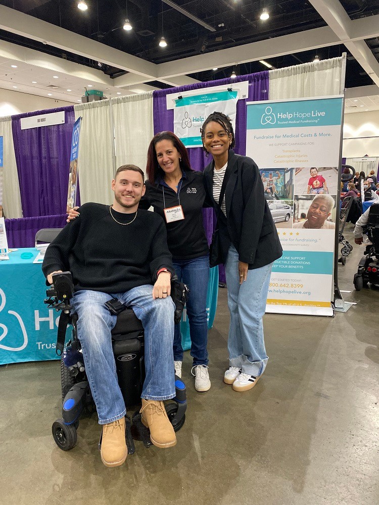 Kelly L Green in front of the Help Hope Live booth and signage at Abilities Expo 2024 with client Cole and his wife Charisma of the YouTube channel Roll with Cole & Charisma. Seated in his power chair, Cole has light skin, short brown hair, a black long-sleeved shirt, jeans, and Timberlands. Kelly has light skin, red and brown straight hair, a black Help Hope Live branded sweater, and jeans. Charisma has brown skin, braided black hair pulled back, a black blazer, and jeans.