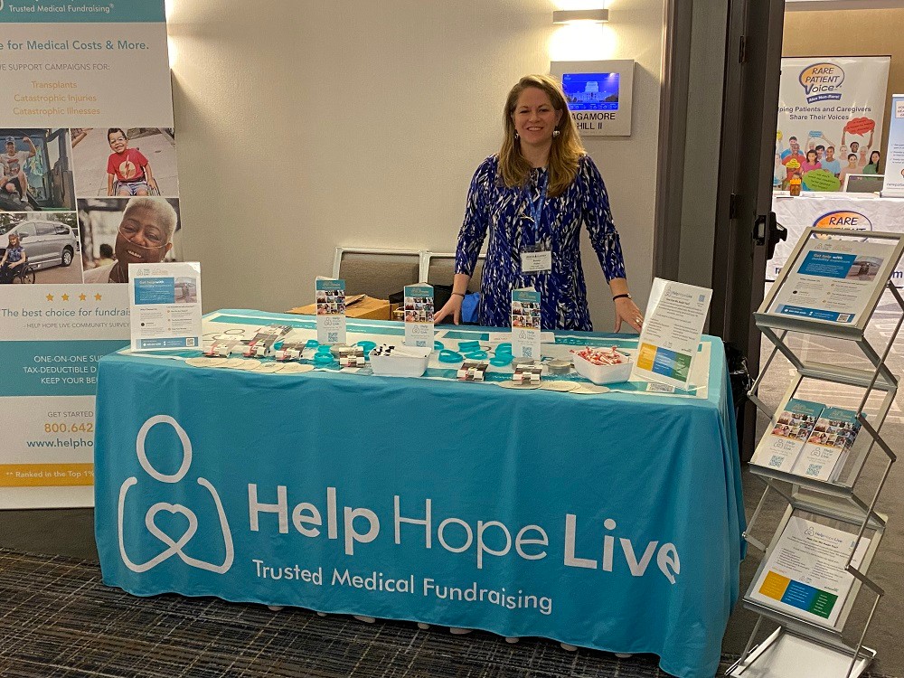 At the Help Hope Live booth at Reeve Summit 2024, which is decked out with teal and the tagline Trusted Medical Fundraising and covered with pens, bracelets, brochures, and materials, Sonny has light skin, golden hair past her shoulders, and a blue and white mosaic patterned dress.