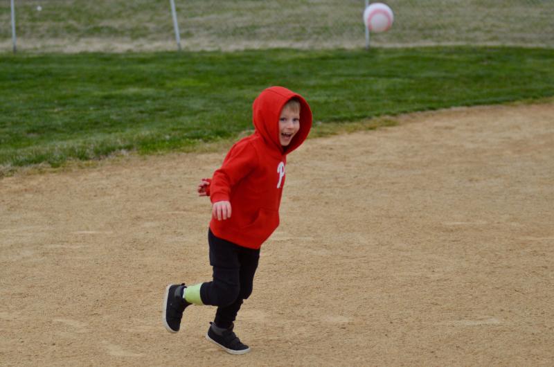 4-year-old Zeke Clark is outdoors on a baseball diamond smiling and running to as field an airborne baseball. He wears a red Phillies logo hoodie and has light skin, short blonde hair, black pants, and a green prosthetic leg on his right side.