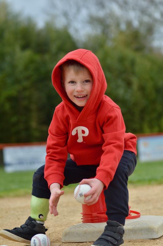 4-year-old Zeke Clark is outdoors on a baseball diamond smiling as he fields two baseballs. He wears a red Phillies logo hoodie and has light skin, short blonde hair, black pants, and a green prosthetic leg on his right side.