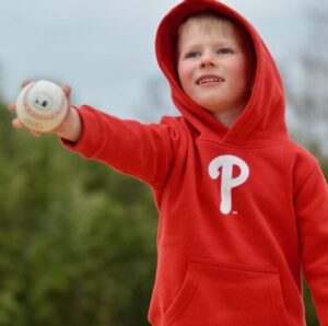 Help Hope Live client Zeke Clark is a young boy with light skin and short blonde hair who is wearing a red hoodie with the Phillies logo and holding a baseball in his right hand.