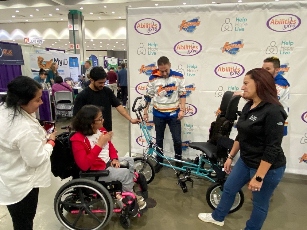 Client Karla Ilhuicatzi is seated in her wheelchair looking at the teal Freedom Concepts adaptive bike she is receiving from Help Hope Live, which sits in front of a step-and-repeat banner featuring logos for Help Hope Live, Abilities Expo, and Freedom Concepts. Karla has light brown skin, shoulder-length brown hair, and glasses. Across from her is Help Hope Live Executive Director Kelly Green, who wears a long-sleeved Help Hope Live black shirt and jeans and has light skin and red-brown straight hair. Two Freedom Concepts staff members wear bright white, orange, and blue branded hoodies. Members of Karla's family are standing near her.