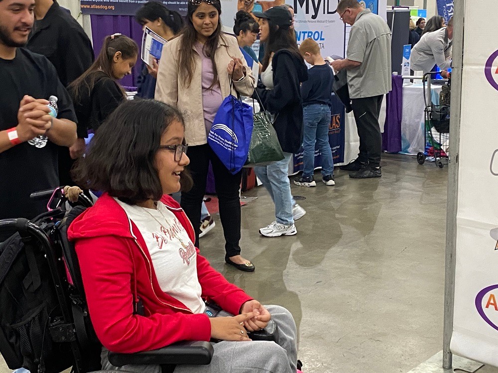 Client Karla Ilhuicatzi is visibly excited as she is seated in her wheelchair looking at the teal Freedom Concepts adaptive bike she is receiving from Help Hope Live, which sits in front of a step-and-repeat banner featuring logos for Help Hope Live, Abilities Expo, and Freedom Concepts. Karla has light brown skin, shoulder-length brown hair, and glasses.