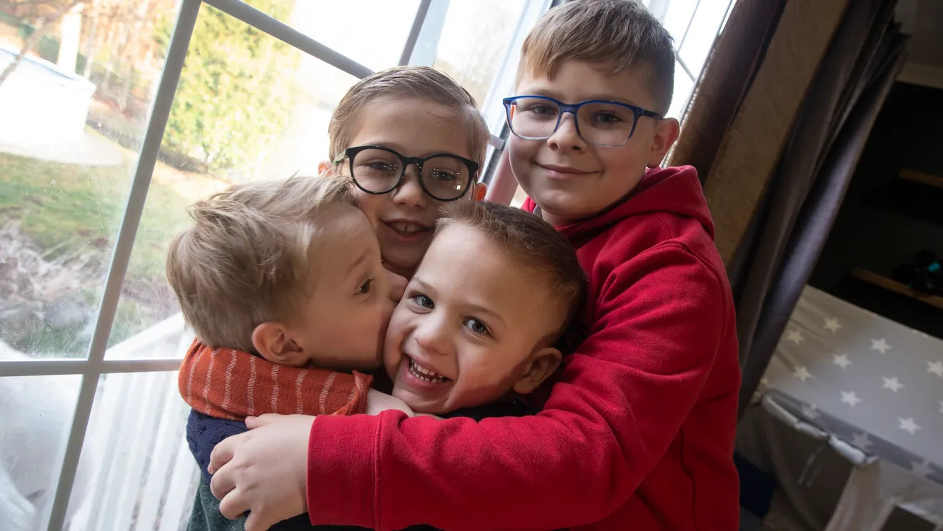 Kids Julian and Ashton Laskowski are embracing two younger kids in a group hug. All four boys have light skin and short sandy blonde hair. The two oldest kids Julian and Ashton have black glasses.