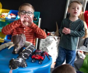 Kids Julian and Ashton Laskowski are in a playroom setting. Both boys have light skin and short blonde hair. One of them has black round glasses and is making a heart shape with his hands above a table with toy cars and dinosaurs.