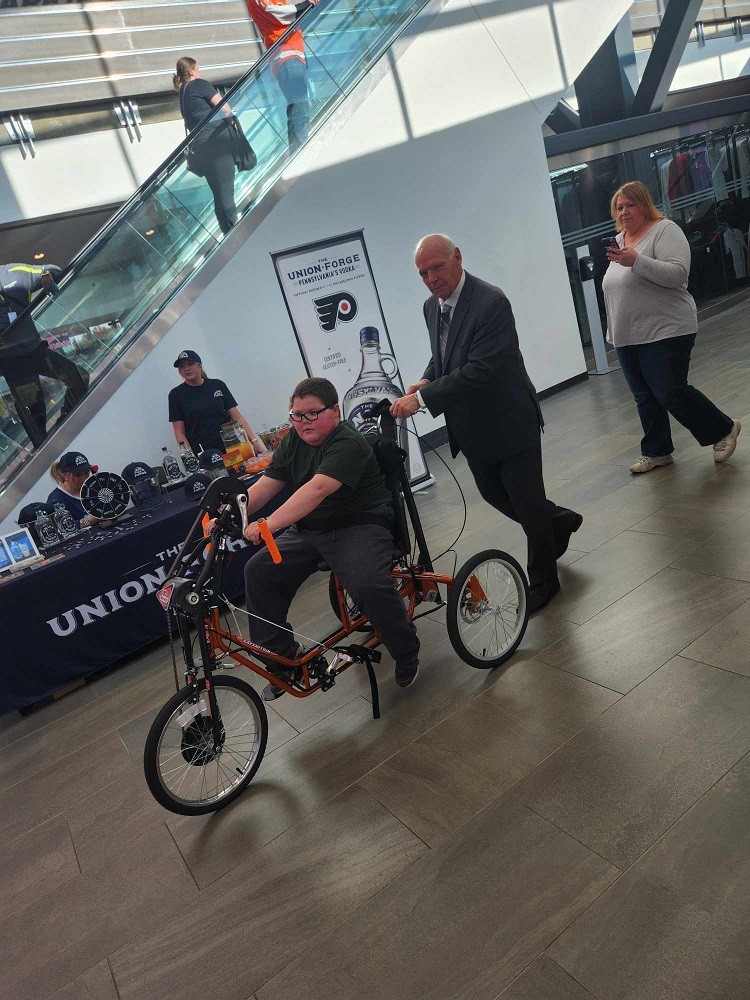 Flyers Alumni Bob Kelly gives Dylan McDonnell a push on his new adaptive bike at Wells Fargo center. Bob has light skin, white hair, and a suit. Dylan has light skin and black glasses and is a teen. The bike is orange and Philadelphia Flyers themed.