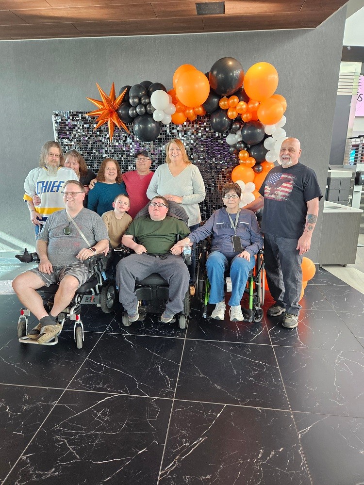 With decorative orange and black Flyers-themed balloons behind them at Wells Fargo center, Dylan McDonnell and his family smile. Dylan is a teen with light skin, brown hair, and black glasses who is seated in a black powerchair. His father is also seated in a power chair.