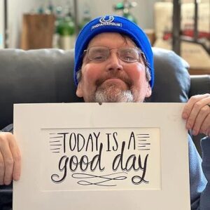 Edward Cardwell is smiling and holding a sign that says Today is a Good Day as he sits in a living room chair. He has light skin, glasses, a gray goatee, and a blue beanie, and he is missing the oxygen tubes he is wearing in most other photos.