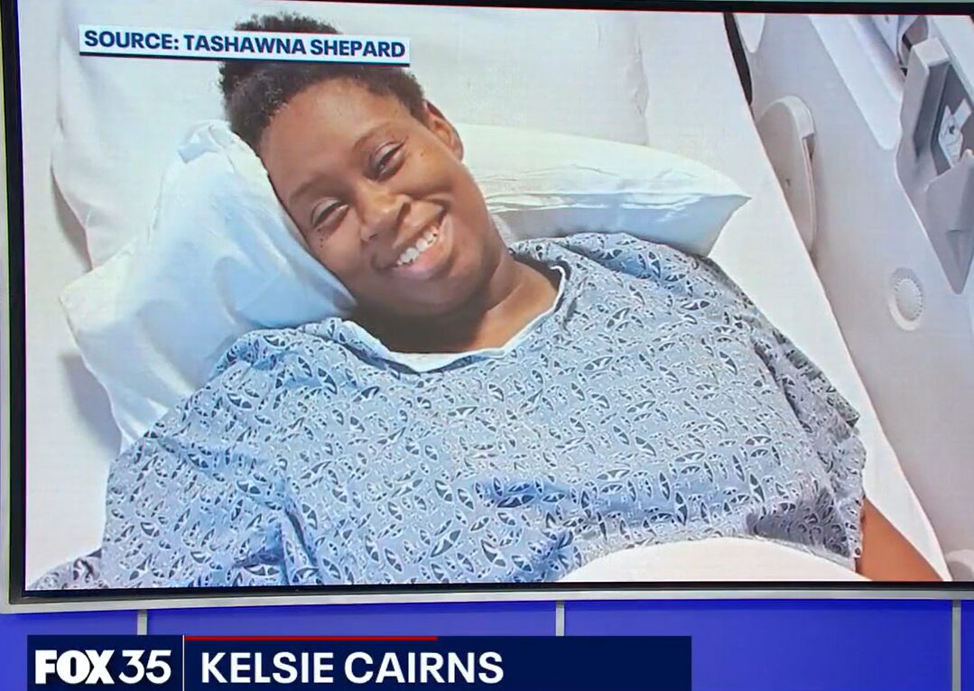 Heart transplant candidate Tashawna Shepard is interviewed by FOX Orlando. The headline reads FOX 35 Kelsie Cairns, which is the name of the reporter. Lying in a hospital bed in a hospital gown, looking tired but with a big smile, Tashawna has brown skin, brown eyes, and black hair pulled up into a bun.