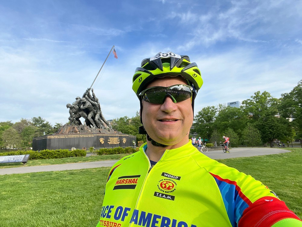 Heart transplant recipient Bill Soloway is at a Faces of America cycling event. He wears bright yellow-green cycling gear with a matching helmet and sunglasses. Behind him in the selfie is a statue of soldiers raising an American flag.