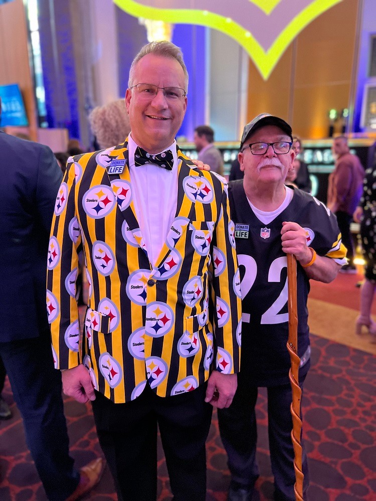 Heart transplant recipient Bill Soloway with his donor's father at a formal event in a casino-like setting. Bill has light skin, short gray hair, and glasses and he wears an eye-popping Steelers themed spots jacket in vivid black and yellow. His donor's father wears a 22 football jersey and a ball cap and has light skin, a white mustache, glasses, and a walking stick in his left hand.