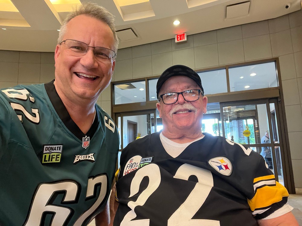 Heart transplant recipient Bill Soloway smiles in a selfie with his donor's father in a lobby setting. Bill has light skin, short gray hair, and a 62 Eagles jersey with a Donate Life patch. His donor's father has light skin, a white mustache, a ball cap, black glasses, and a number 22 Steelers jersey.